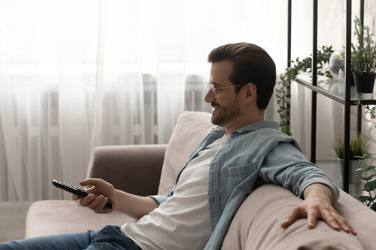 Good Time By Tv Set. Side View Of Relaxed Young Man Sitting On Sofa Watching Tv, Choosing Cable Channel Using Remote Control. Peaceful Guy Selecting Television Programs Spending Evening At Home Alone