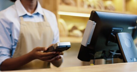 Close up of African American woman seller selling in bakery shop using device to make payment. Caucasian client hand paying with credit card while buying in bakehouse. Business concept