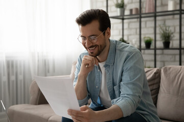 Pleasure to receive good news. Glad young man sitting on sofa reading paper letter of getting reward from corporate office. Smiling businessman hold profitable contract hardcopy enjoying perfect work - obrazy, fototapety, plakaty