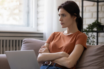 That is a question. Pensive millennial lady sit on couch at home using laptop planning remote work, Thoughtful female distracted from pc screen holding hands crossed. Young woman thinking on problem