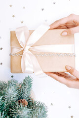 Woman holding a present in a craft cover with beige bow on a white background with decorative stars, branches of spruce.