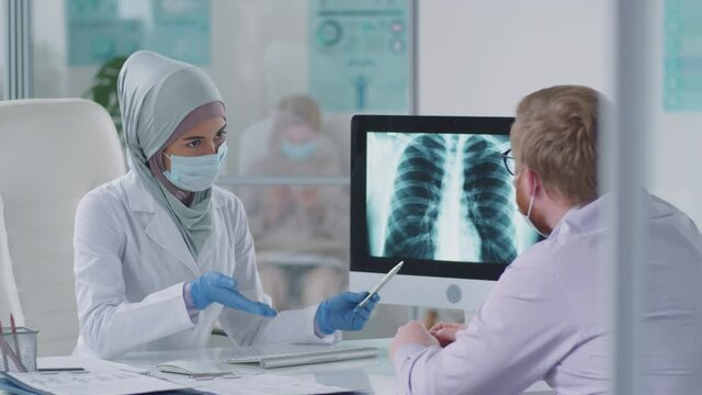 Muslim female doctor in hijab and disposable face mask showing chest x-ray on computer to male patient while giving consultation in medical office during covid-19 pandemic