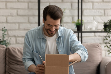 What is inside. Curious millennial male sitting on couch at home opening small cardboard box...