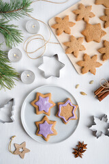 Preparation of sweet savory ginger cookies or gingerbread with violet icing cooked for christmas at winter on white wooden table with cutters, cinnamon, fir tree and candle lights. Vertical, flat lay
