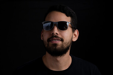 Obraz premium Young bearded man headshot over black background. He is smiling and is wearing sunglasses.