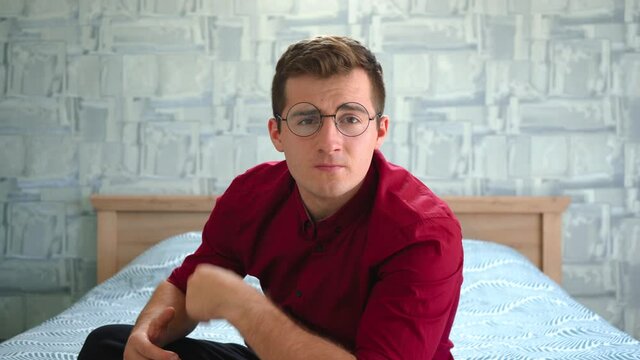 a man in round glasses sits on the bed and looks at the camera