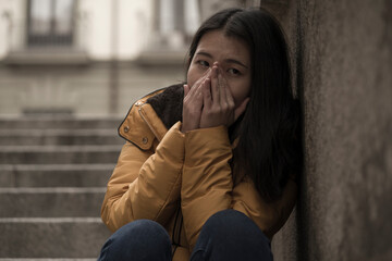 young attractive sad and depressed Chinese woman in winter jacket sitting outdoors on street corner...