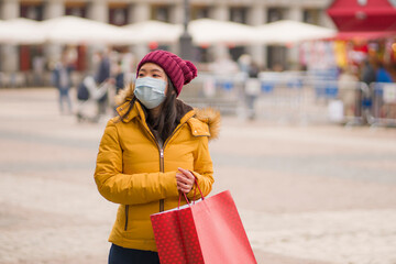 Asian girl enjoying Christmas shopping during covid19 - young happy and beautiful Japanese woman with mask holding red shopping bag buying presents on xmas street market
