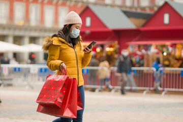 Asian girl enjoying Christmas shopping during covid19 - young happy and beautiful Japanese woman with mask holding red shopping bag buying presents on xmas street market