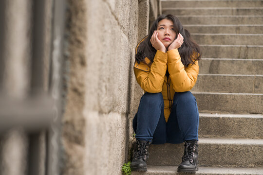 dramatic lifestyle portrait of young attractive sad and depressed Japanese woman in winter jacket sitting outdoors on street corner staircase suffering depression problem feeling helpless
