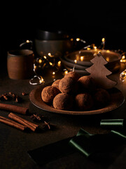 Chocolate truffles in cocoa dusting on dark brown ceramic plate decorated with wooden fir. Ceramic plates, cups, Christmas lights behind it. Cinnamon sticks, star anise and decorative ribbon nearby