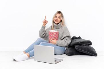 Teenager blonde girl eating popcorn while watching a movie on the laptop showing and lifting a finger in sign of the best