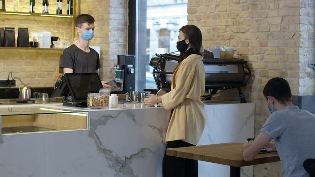 Young Caucasian Brunette Woman In Covid Face Mask Paying For Takeaway Coffee In Cafe And Leaving. Barista Serving Client Indoors. New Normal Coronavirus Lifestyle Cinema 4k ProRes HQ.