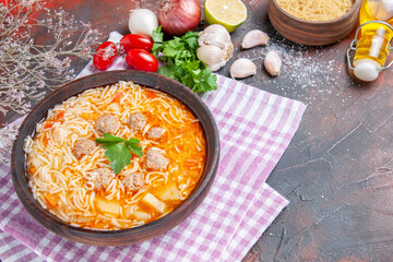 Side view of delicious chicken soup with noodles greens and spoon on pink stripped towel oil bottle garlic tomatoes lemon on dark background