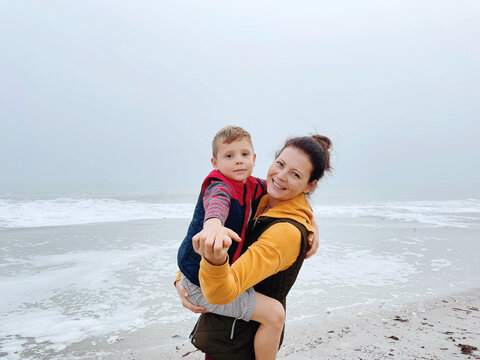 Mother and son walk along the foggy beach and take a photo on the phone. Portrait of happy mom and child boy on the background of cloudy weather made with phone. Family time together. 