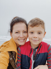 Mother and son walk along the foggy beach and take a selfie on the phone. Portrait of happy mom kiss her child boy on the background of cloudy weather made with phone. Family time together. 