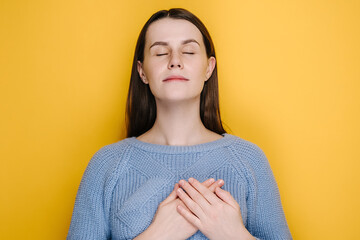 Smiling millennial young girl put folded palms on heart, isolated on yellow studio background,...