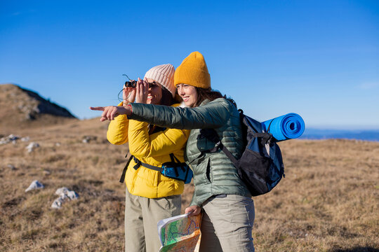 Two female friends are hiking at the top of the mountain. They are using a geographical map and binoculars.