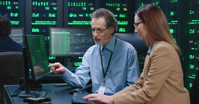 Senior employee of system administrator with glasses sits at table with female intern programmer and explains to her workflows showing on computer screen.