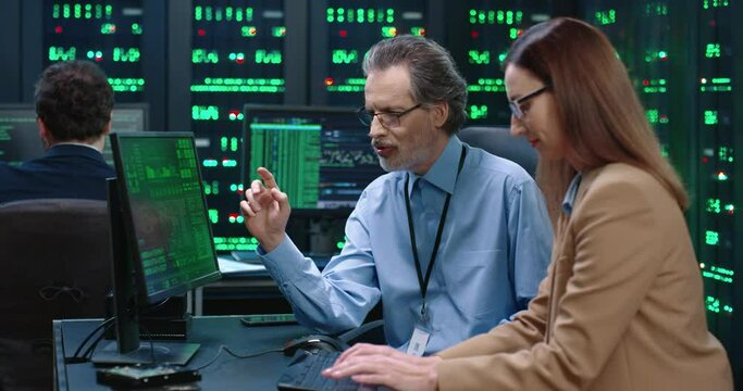 Senior employee of system administrator in glasses sits at table with female programmer and gives her instructions on how to perform work task.