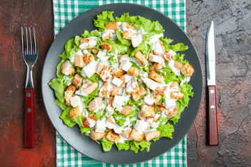 top view caesar salad on oval plate green white checkered tablecloth fork and knife on dark red background