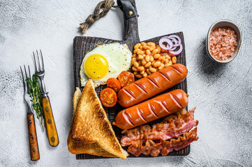 Full fry up English breakfastt with fried eggs, sausages, bacon, beans and toasts on a wooden cutting board. White background. top view