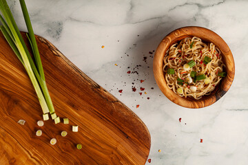 A wooden bowl of cooked ramen noodles seasoned with herbs and freshly chopped spring onions. The bowl is served on marble surface with the chopping board near by. Spices are seen on marble