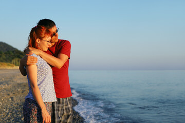 Young couple in love stand on the seashore in the evening and look into the distance in appeasement in an embrace. Valentines day concept with copy space