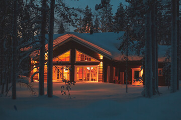 A night view of cozy wooden scandinavian cabin cottage chalet house covered in snow near ski resort in winter with the lights turn on, evening picture