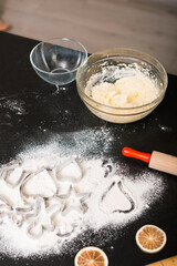 Vertical photo of a cooking table with flour, cookies cutting shapes and a bowl with dough.