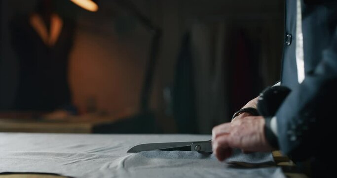 Cinematic shot of experienced tailor is cutting with scissors fine quality fabric for creation custom haute couture apparel in tailoring atelier. Concept of handmade, couturier and tradition.