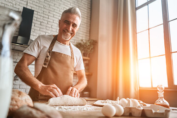 Smiling adult male wearing apron and cooking in the kitchen at home. Happy mature man preparing the dough while kneading the dough on a wooden counter with flour and a roll pin. Baking concept