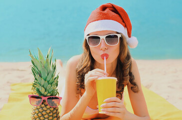 Christmas portrait of happy young woman drinking fresh juice in red santa hat with pineapple lying...