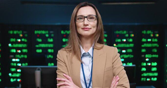 Close up portrait of female system administrator programmer in jacket looking at camera and smiling with arms crossed standing in office on background of running servers.