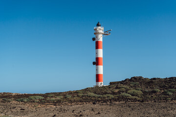 An old lighthouse with white and red stripes on the shore of a rocky island. Clear sunny blue sky.