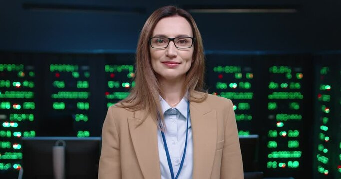 Close up portrait of female system administrator programmer, turning head and looking at camera and smiling, standing in office on background of running servers.