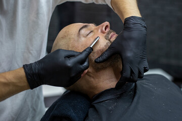 Hands of a barber in black gloves shave the beard of a client with a straight razor in the barber...