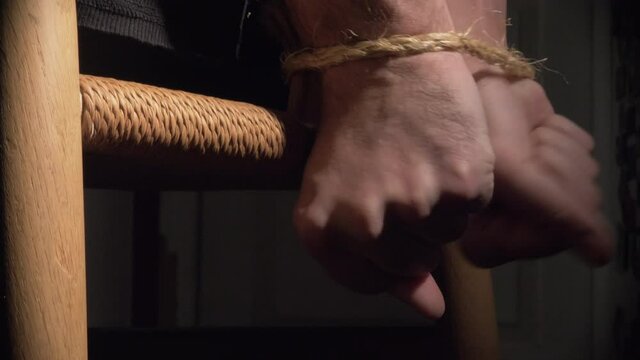 Man Struggles With Hands Tied Behind Back To Wooden Chair Inside A Dark Room With Spotlight Close Up