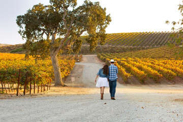 Woman in white dress and denim jacket and Man with cowboy hat and plaid shirt walking as a couple...