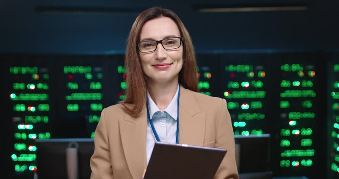 Portrait of woman system administrator programmer looking at tablet raising head and smiling at camera standing in office against background of running servers.