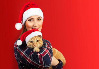 Cute smiling beautiful girl in Christmas hat hugging her cat in Christmas hat too on red background.