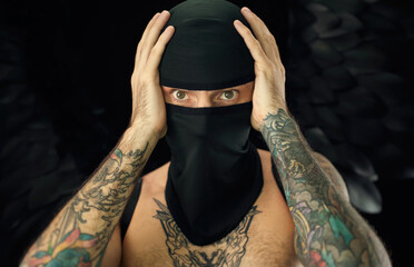 portrait of a guy in a balaclava with a bare torso in tattoos holding his head