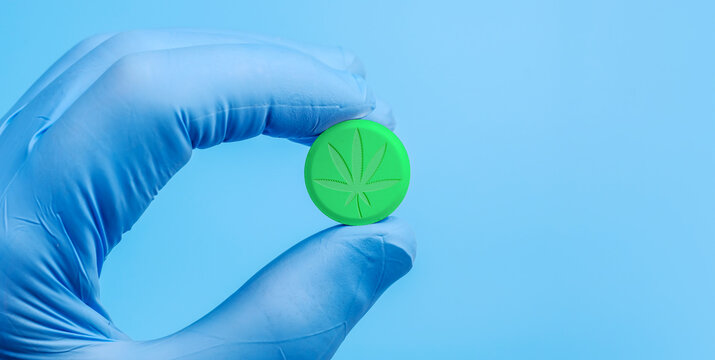 Big green pill with embossed image of marijuana leaf in hand in a medical glove on blue background. Legalization of soft drugs and use in medicine. Close-up view, copy space