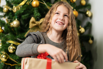 Front view portrait of small caucasian girl eight years old sitting in front of the christmas tree at home with wrapped box gift presents looking to the camera joyful