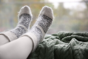 Woman wearing warm socks on knitted plaid near window, closeup. Space for text