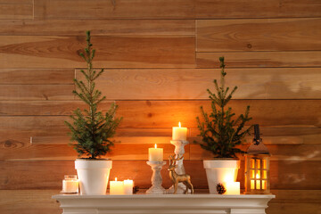 Fototapeta na wymiar Small potted firs, candles and decor elements on white mantel near wooden wall