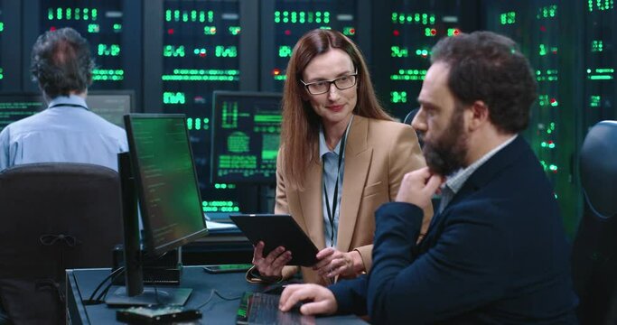 Two programmers work together in server room at desktop: woman holds tablet, man enters code on computer keyboard.