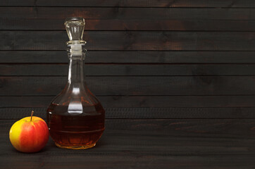 A decanter with apple cider vinegar and whole red yellow apple on dark wooden background.