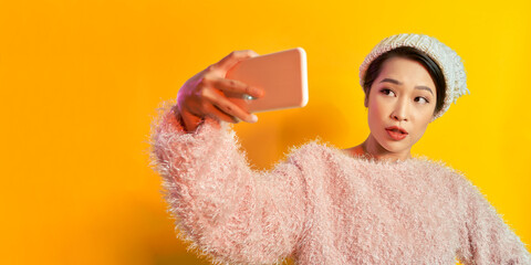 Lovely beautiful asian woman in winter clothes taking selfie, isolated on yellow background.