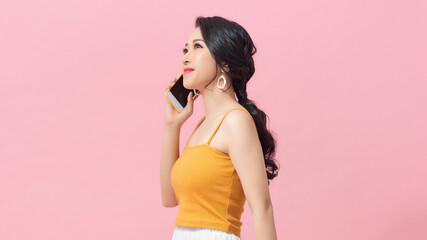 Happy asian woman in casual clothes talking by smartphone and looking away over pink background